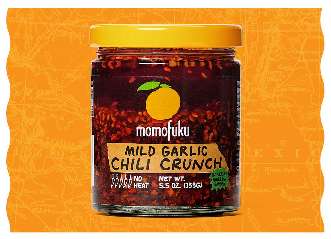 A jar of Momofuku Mild Garlic Chili Crunch with a yellow lid against an orange, map-like background.