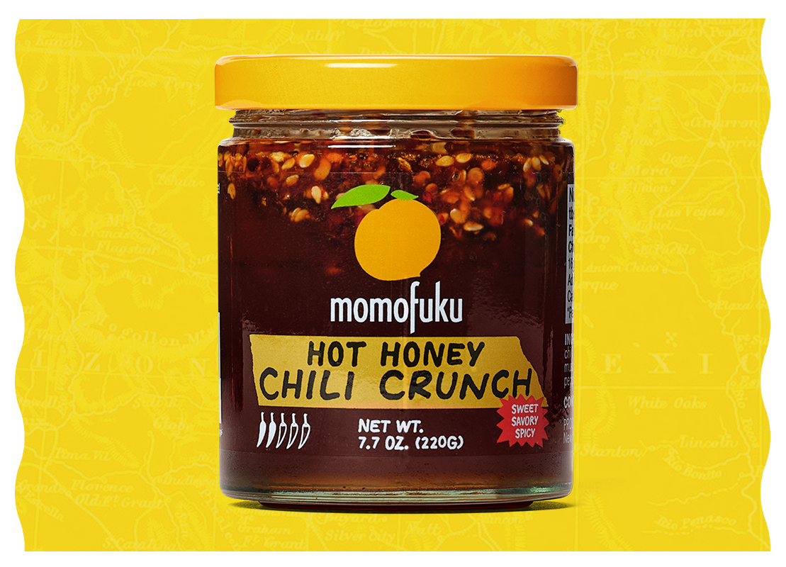 A jar of Momofuku Hot Honey Chili Crunch set against a bright yellow illustrated background.
