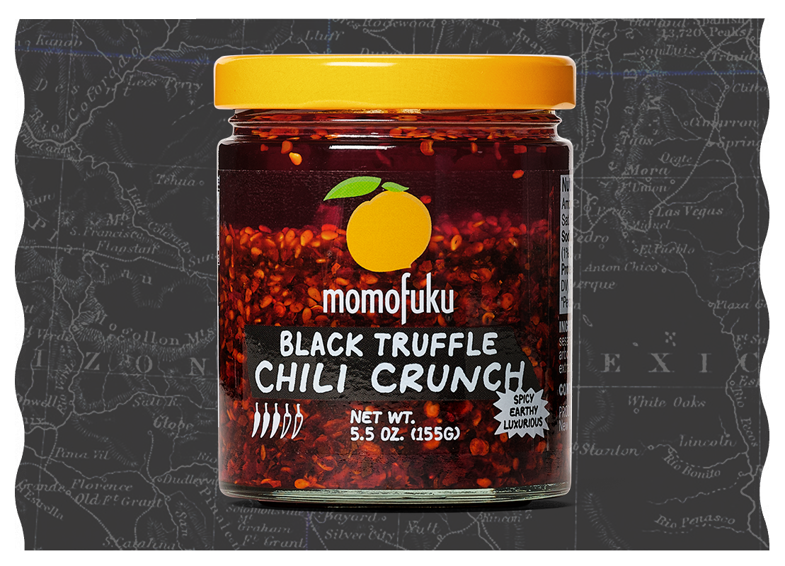 A glass jar of Momofuku Black Truffle Chili Crunch with a yellow lid, set against a dark map.