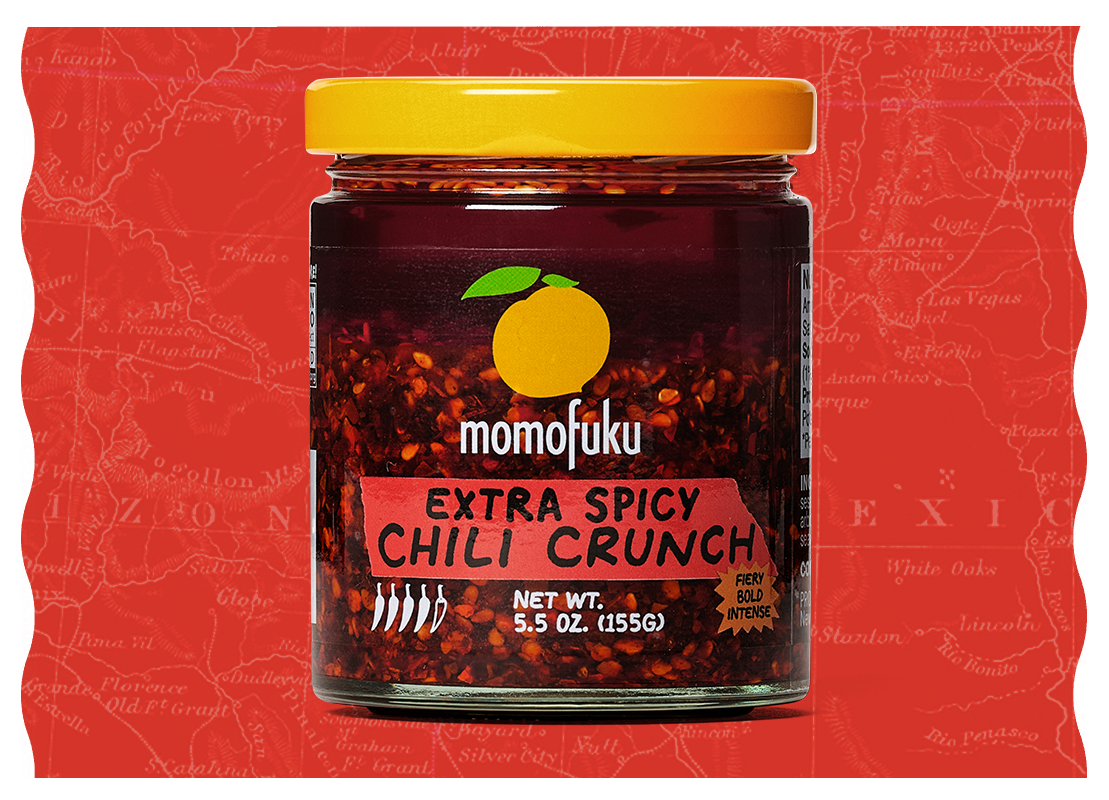 A glass jar of Momofuku Extra Spicy Chili Crunch with a yellow lid, against a red background.