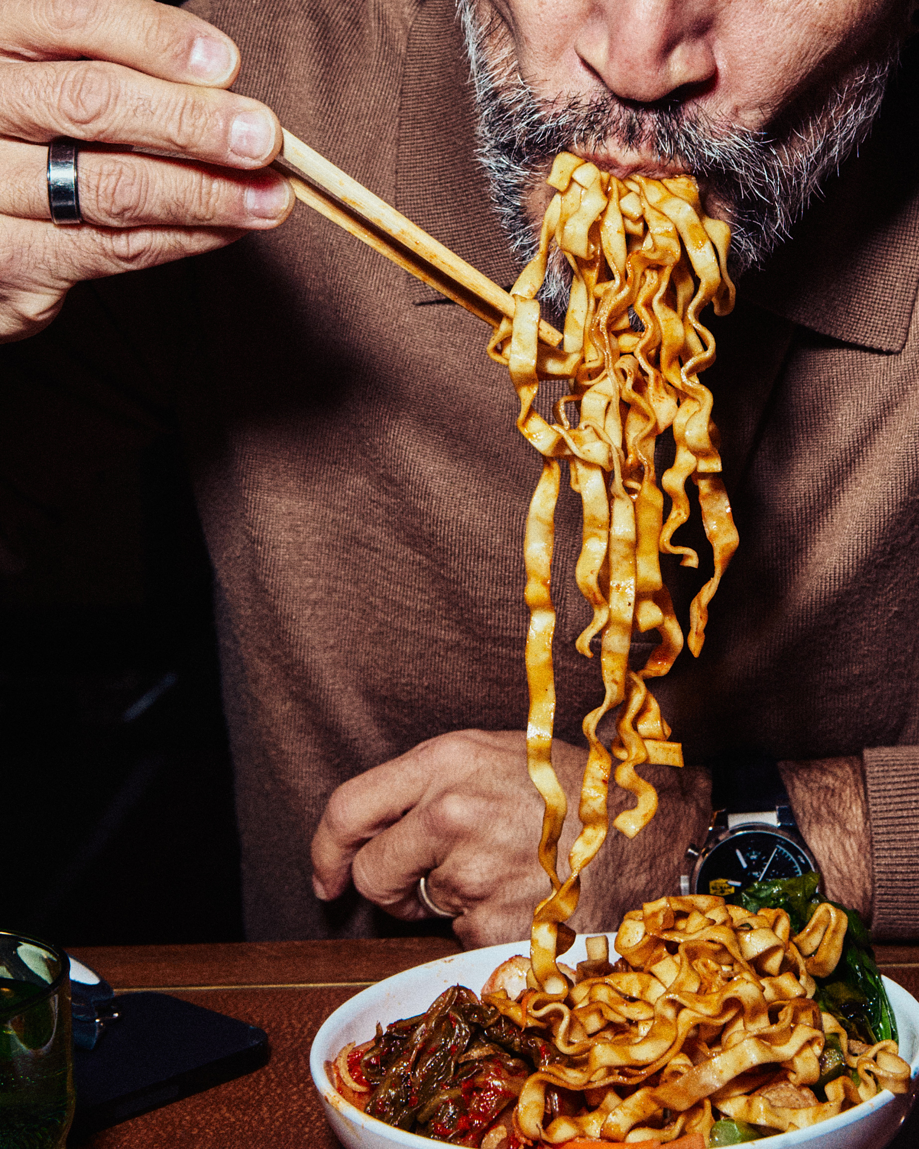A close-up of a bearded man eating a large mouthful of wavy noodles with chopsticks.