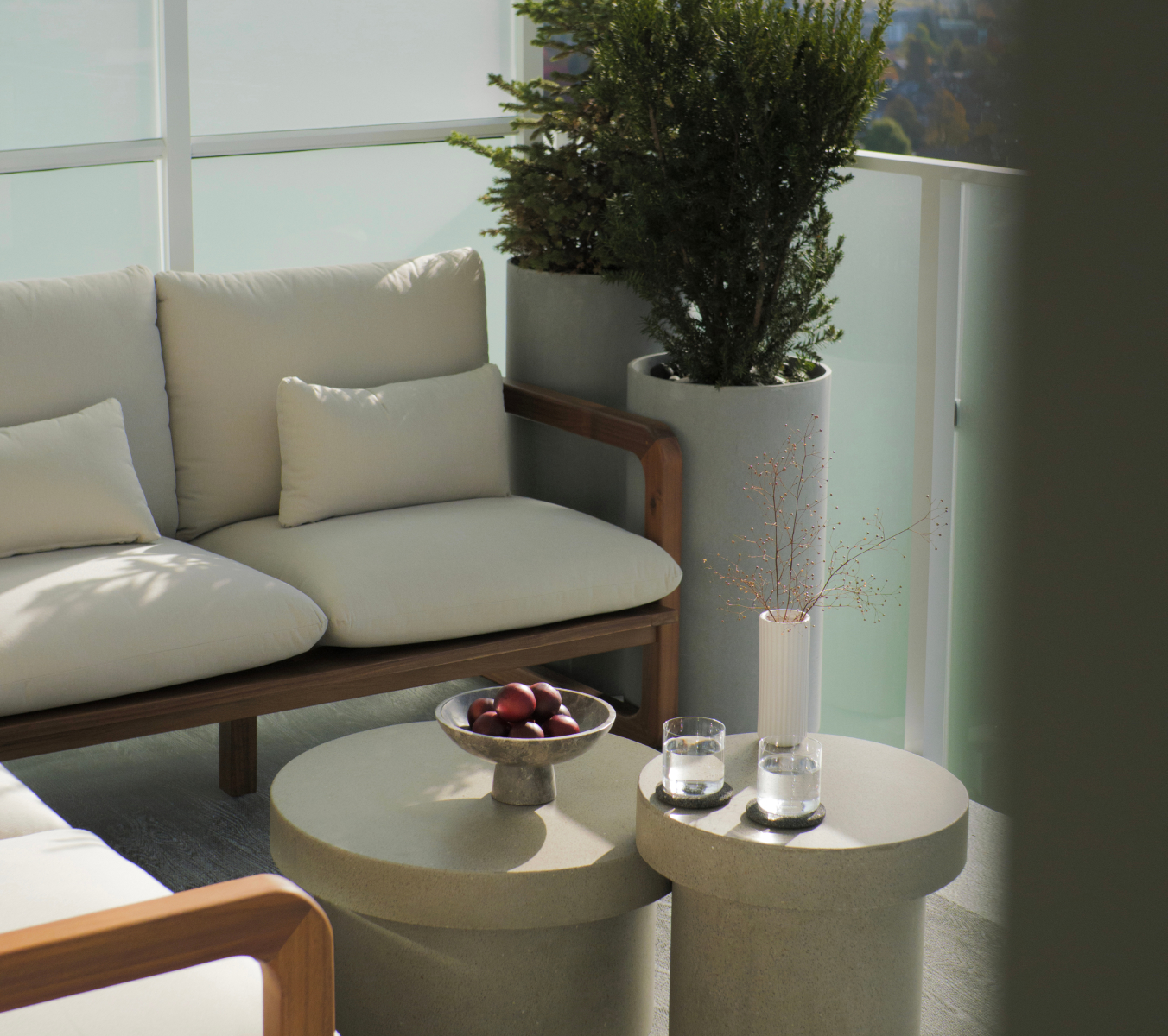 Outdoor seating area with beige cushions, potted plants, and round tables with glasses and a fruit bowl.