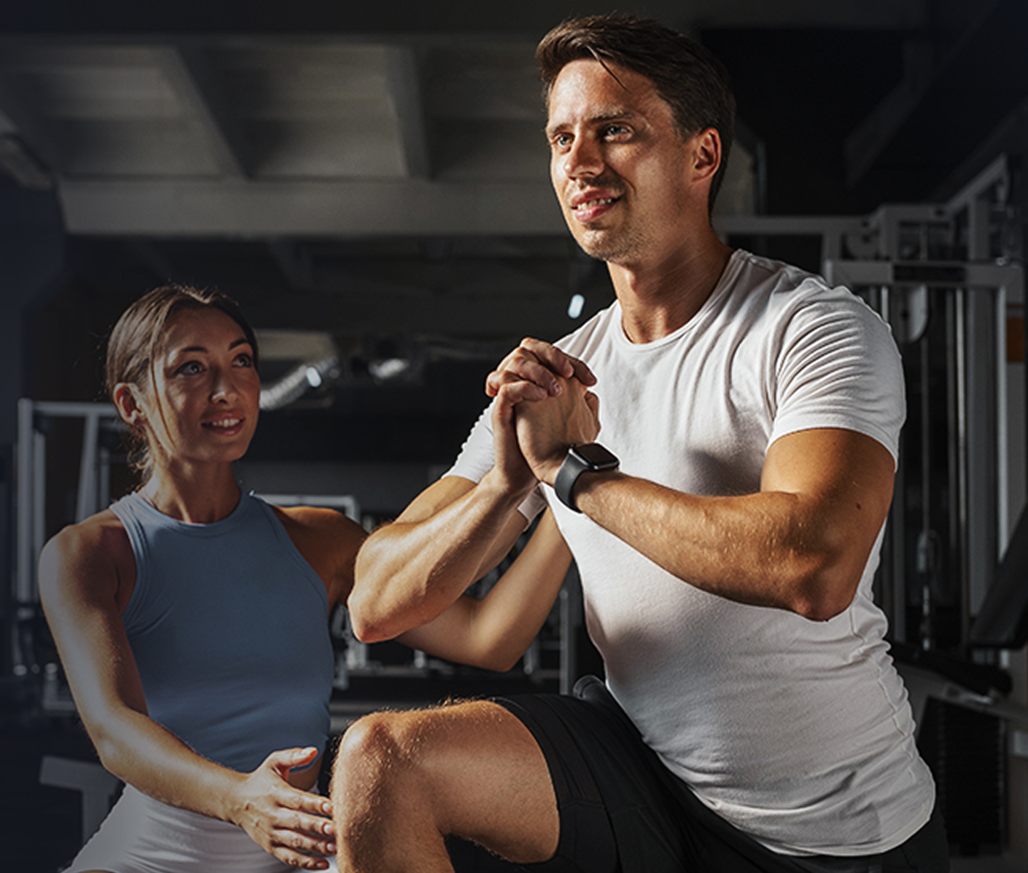 Two men in a gym, one lifting dumbbells, the other assisting.