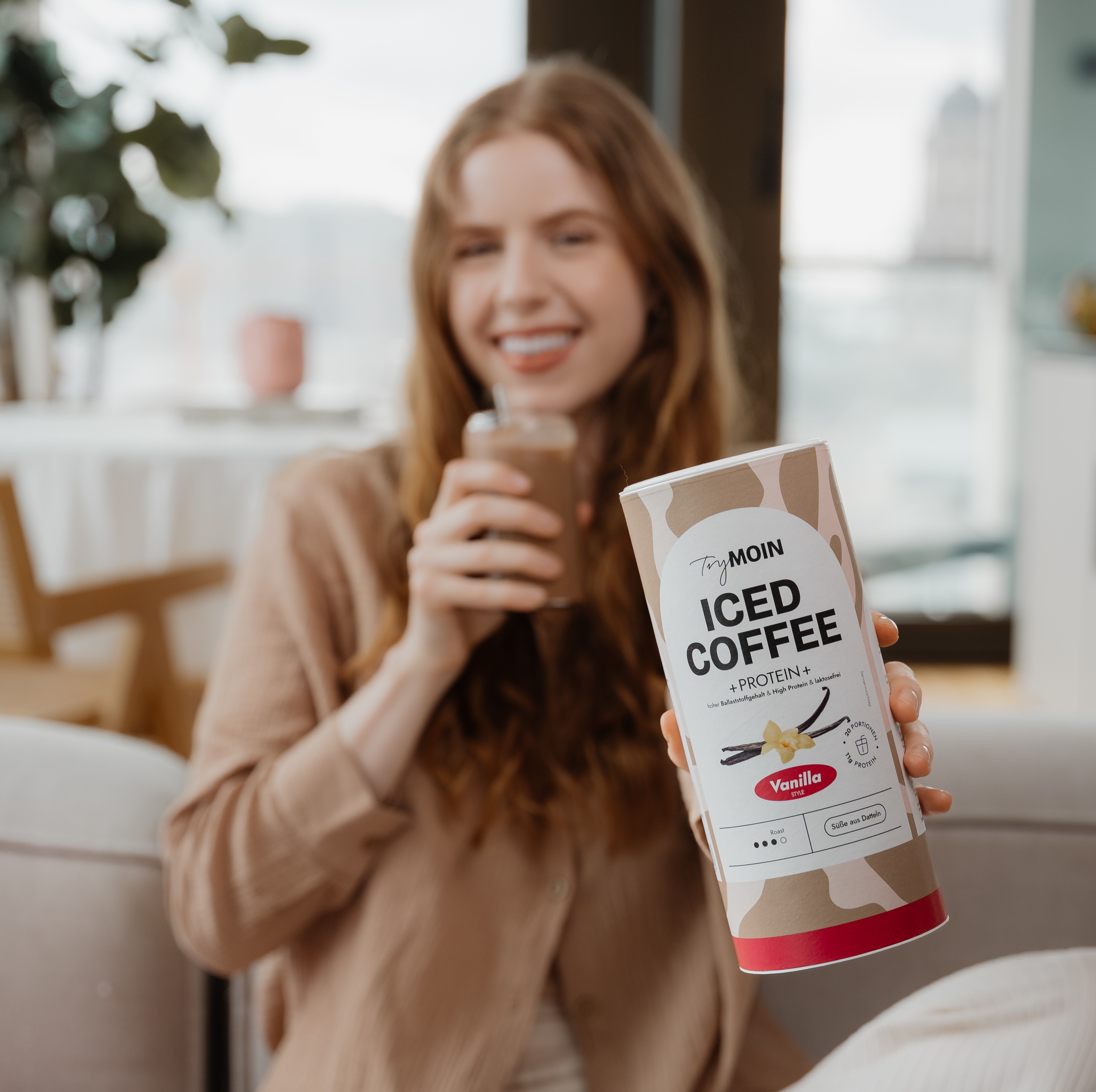 Person holding a can of vanilla iced coffee, smiling in a cozy room.
