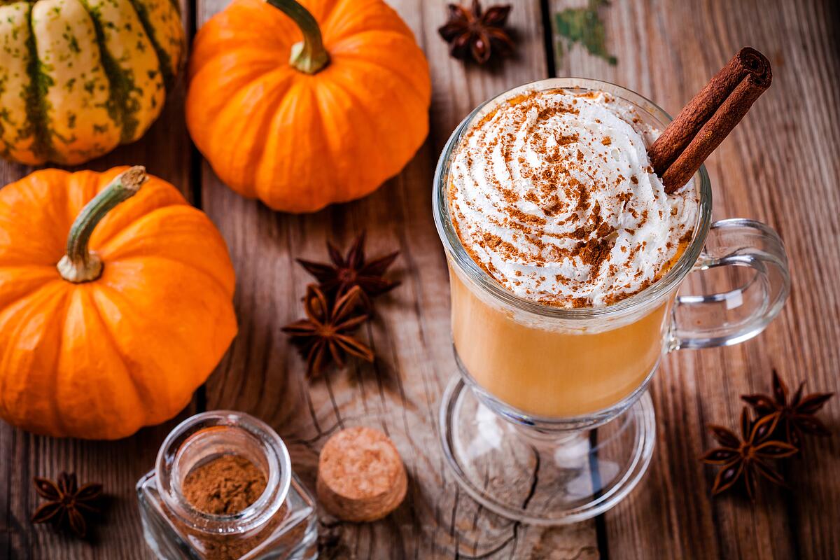 Pumpkin spice latte with whipped cream and cinnamon stick, surrounded by pumpkins and spices.