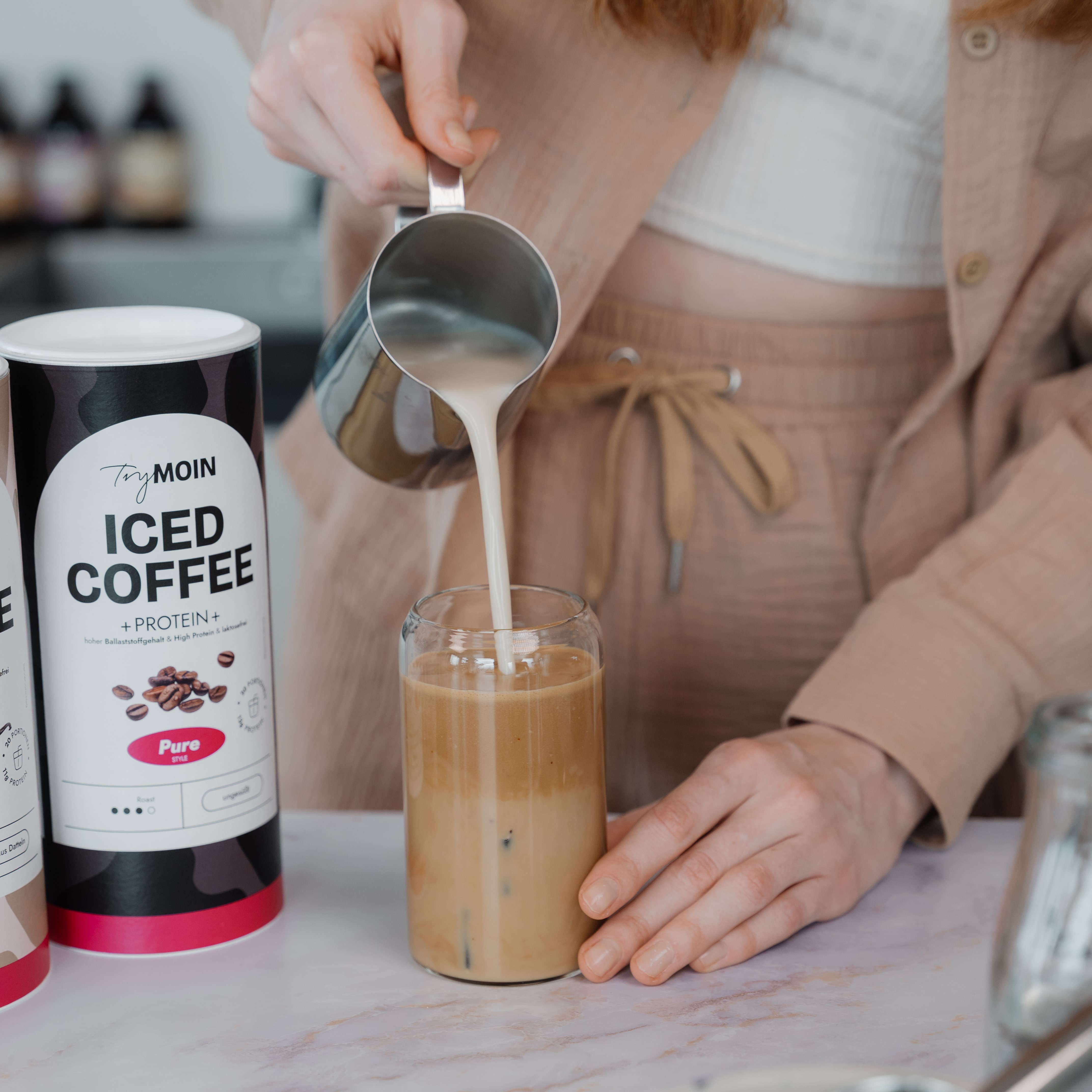 Person pouring milk into a glass of iced coffee on a table.