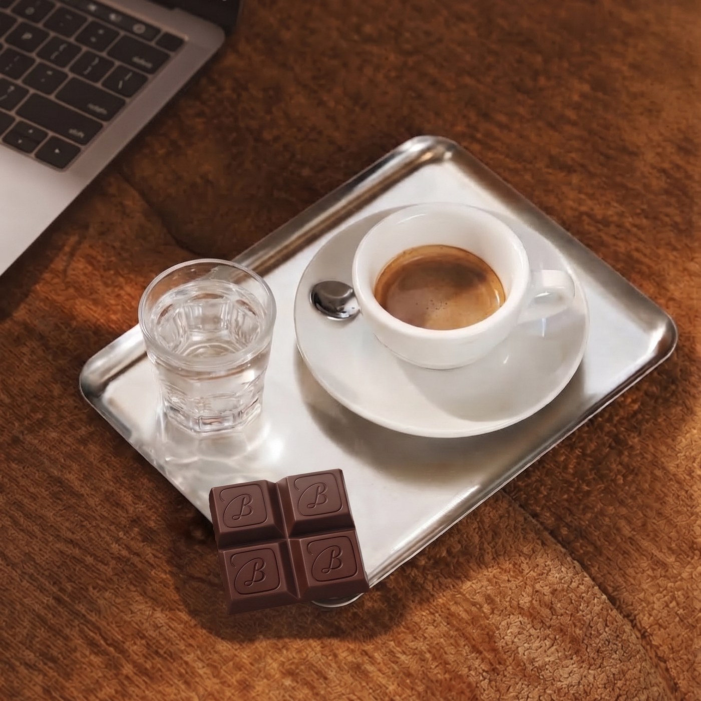 An espresso, glass of water, and chocolate on a silver tray next to a laptop.