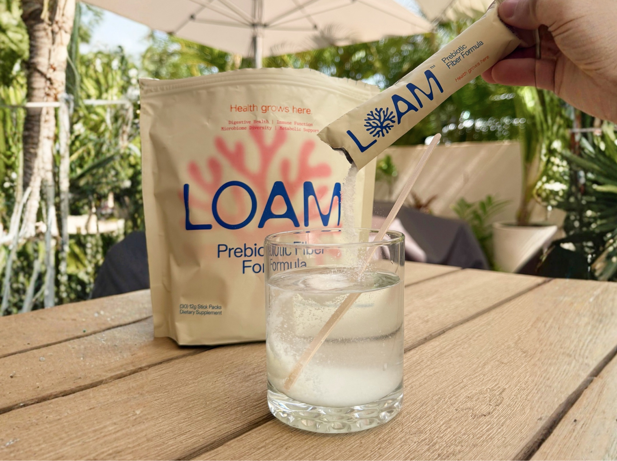 A hand pours a packet of Loam Prebiotic Fiber Formula into a glass of water on a table.