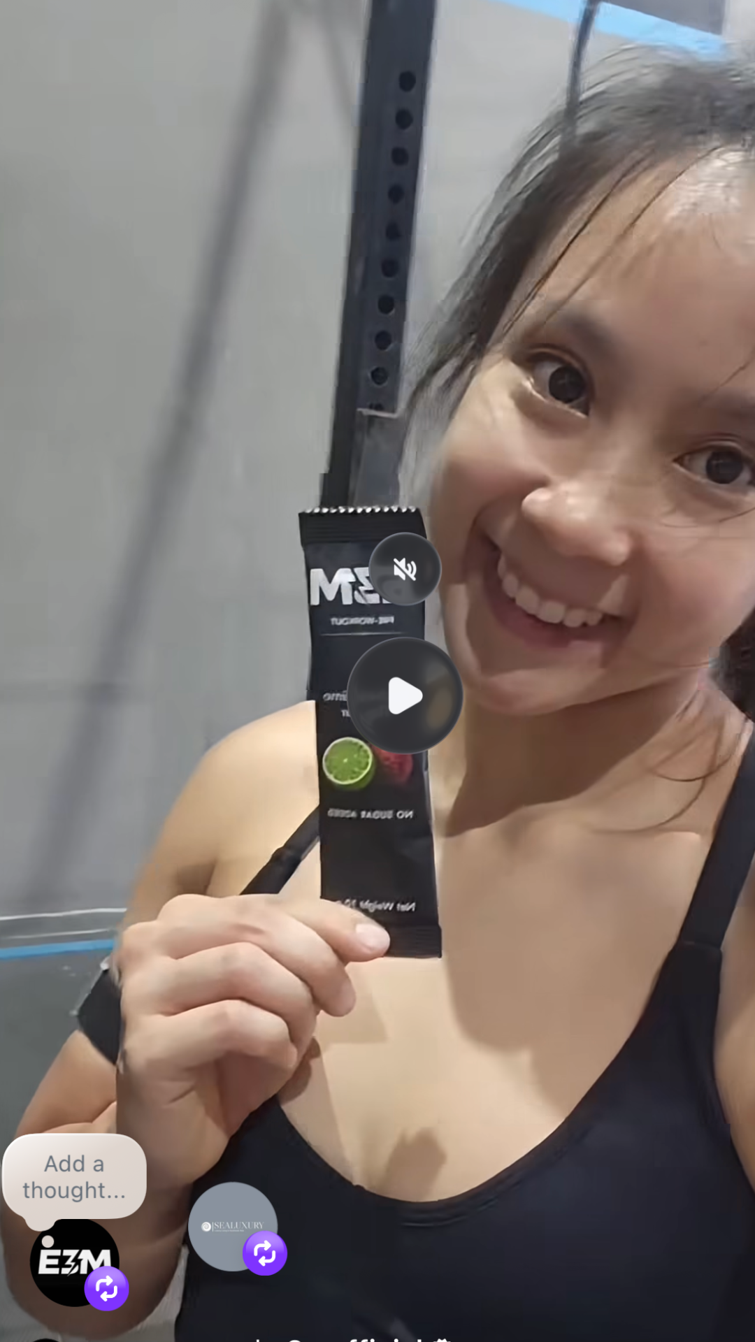 A smiling woman in a gym holds up a black supplement packet.