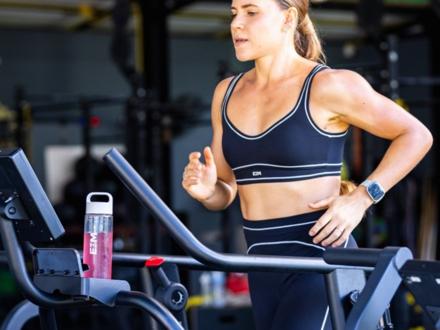 A woman in a black sports bra and leggings runs on a treadmill inside a gym.