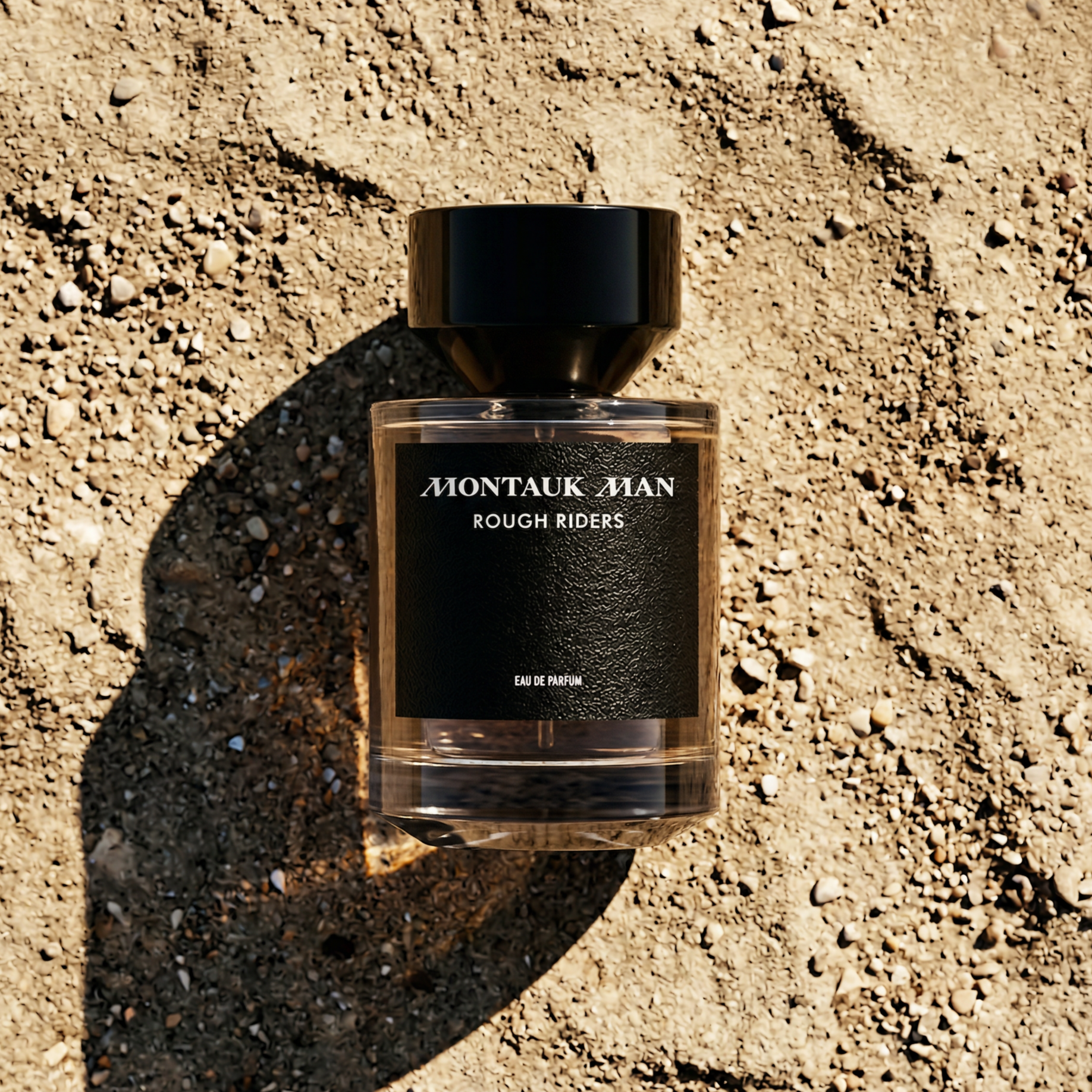 An overhead shot of a bottle of Montauk Man Rough Riders perfume on sandy, pebbly ground.