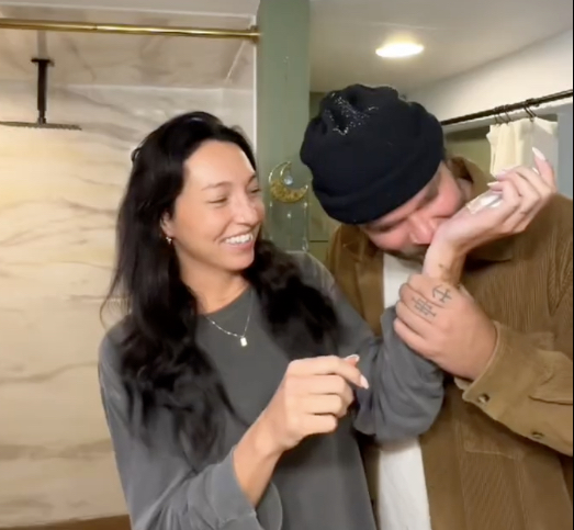 Two people in a bathroom, one smelling the other's wrist, both smiling.