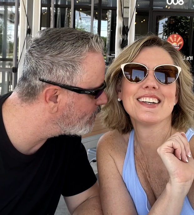 Two people sitting outside, one smiling wearing sunglasses, the other leaning in, wearing black shirt.
