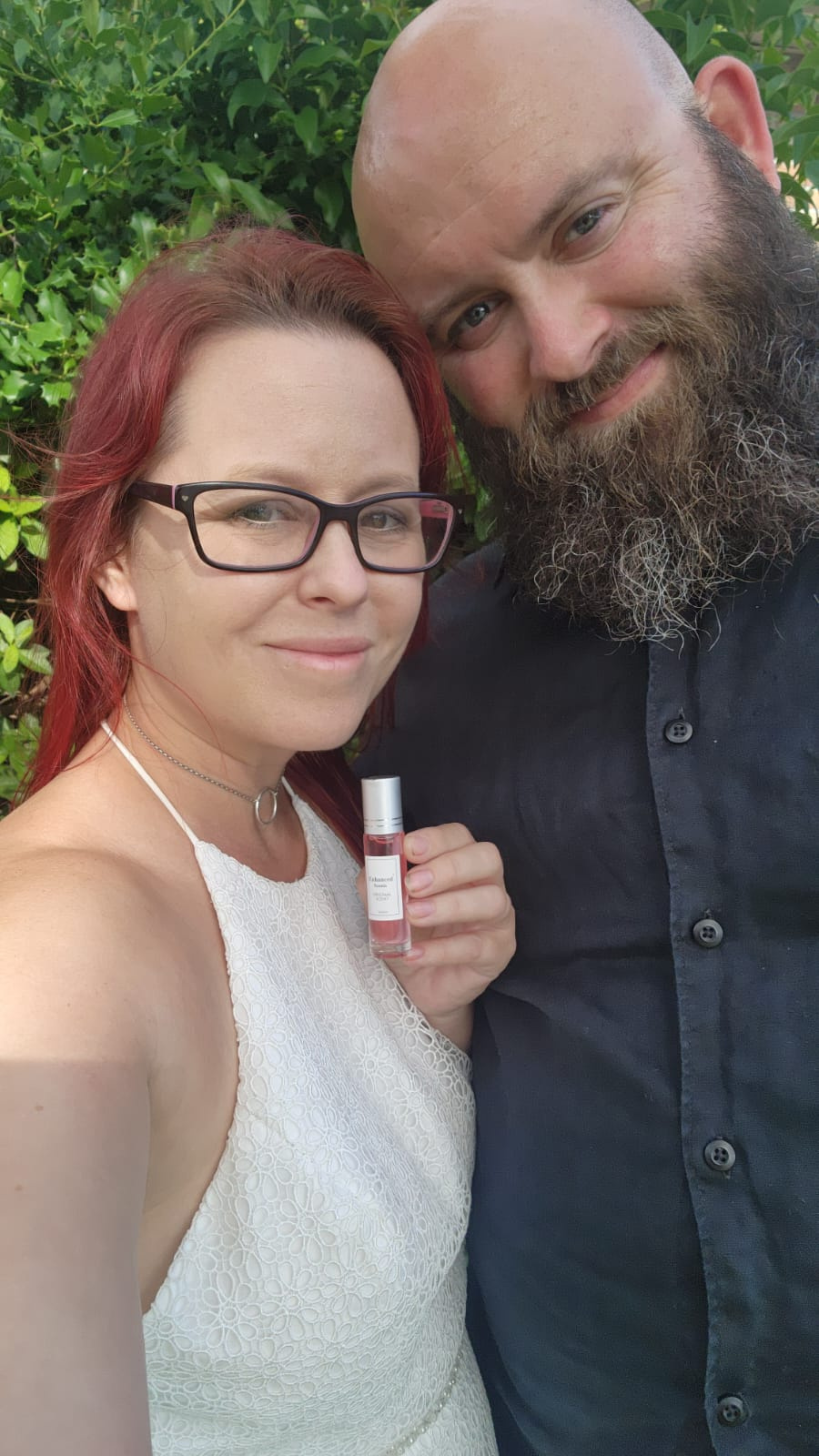 Two people smiling, one holding a small bottle, with greenery in the background.