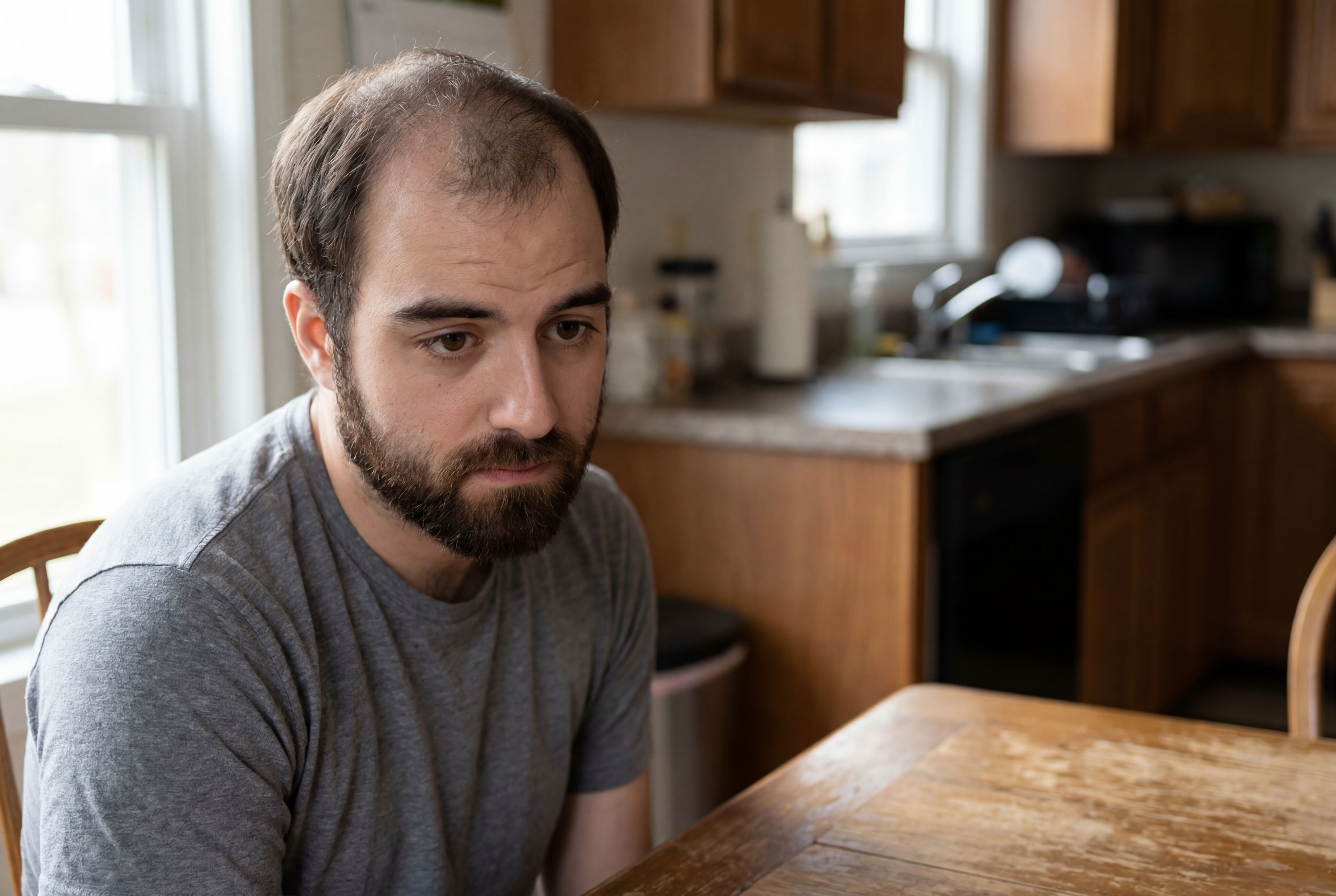 A man with a beard and thinning hair, wearing a gray t-shirt, sits at a wooden kitchen table.