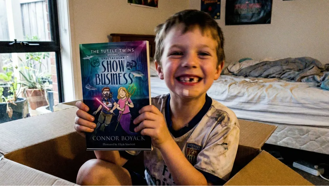 A smiling boy with a missing tooth sits in a cardboard box holding up a Tuttle Twins book.