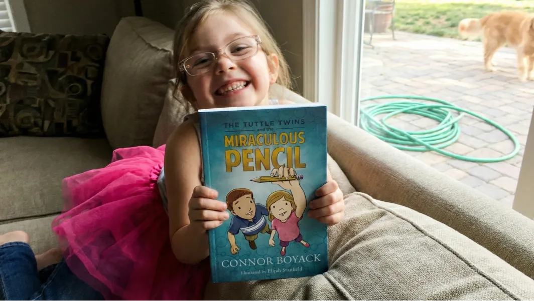 A smiling young girl with glasses holds up a copy of 'The Tuttle Twins and the Miraculous Pencil'.
