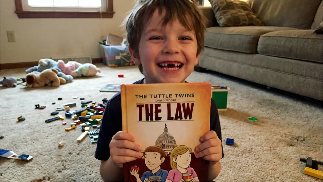 A smiling young boy with a missing tooth holds up a copy of 'The Tuttle Twins Learn About The Law'.