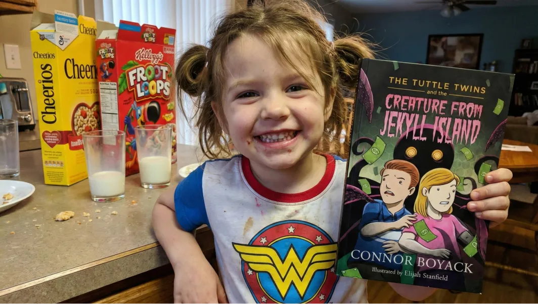 A smiling young girl in a Wonder Woman shirt holds up a book next to boxes of cereal.
