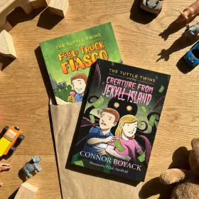 An overhead view of two 'Tuttle Twins' books, 'Food Truck Fiasco' and 'Creature from Jekyll Island,' surrounded by toys.