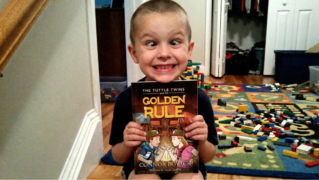 A young boy with a buzz cut makes a silly cross-eyed face while holding a book.
