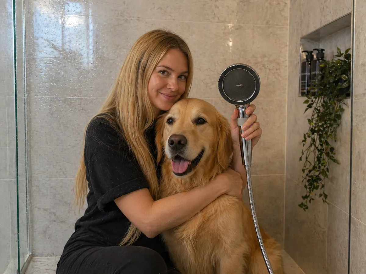 A woman with long blonde hair hugs a golden retriever in a tiled shower, holding a shower head.
