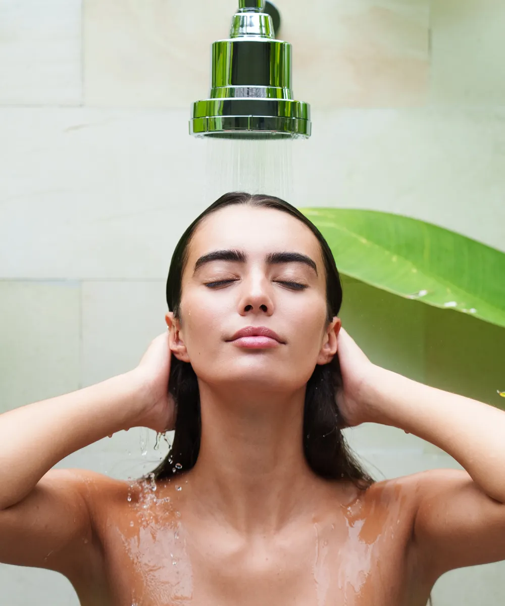 A woman with her eyes closed stands under a running shower with her hands behind her head.