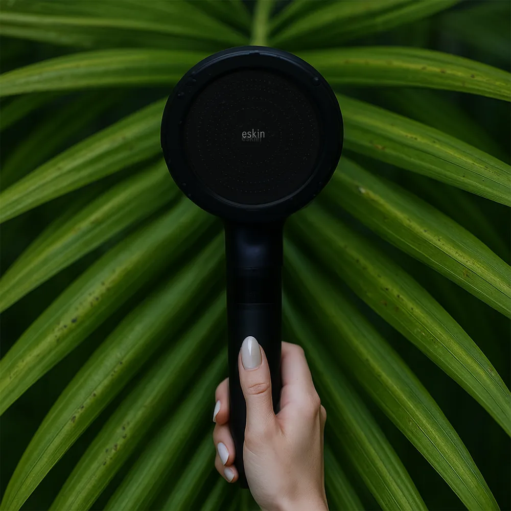 Hand holding a black showerhead against green leaves background.
