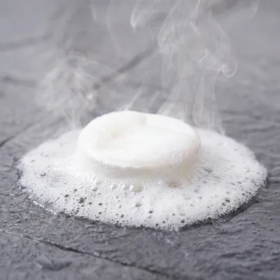 A close-up of a white, circular tablet fizzing and smoking on a dark, wet stone surface.
