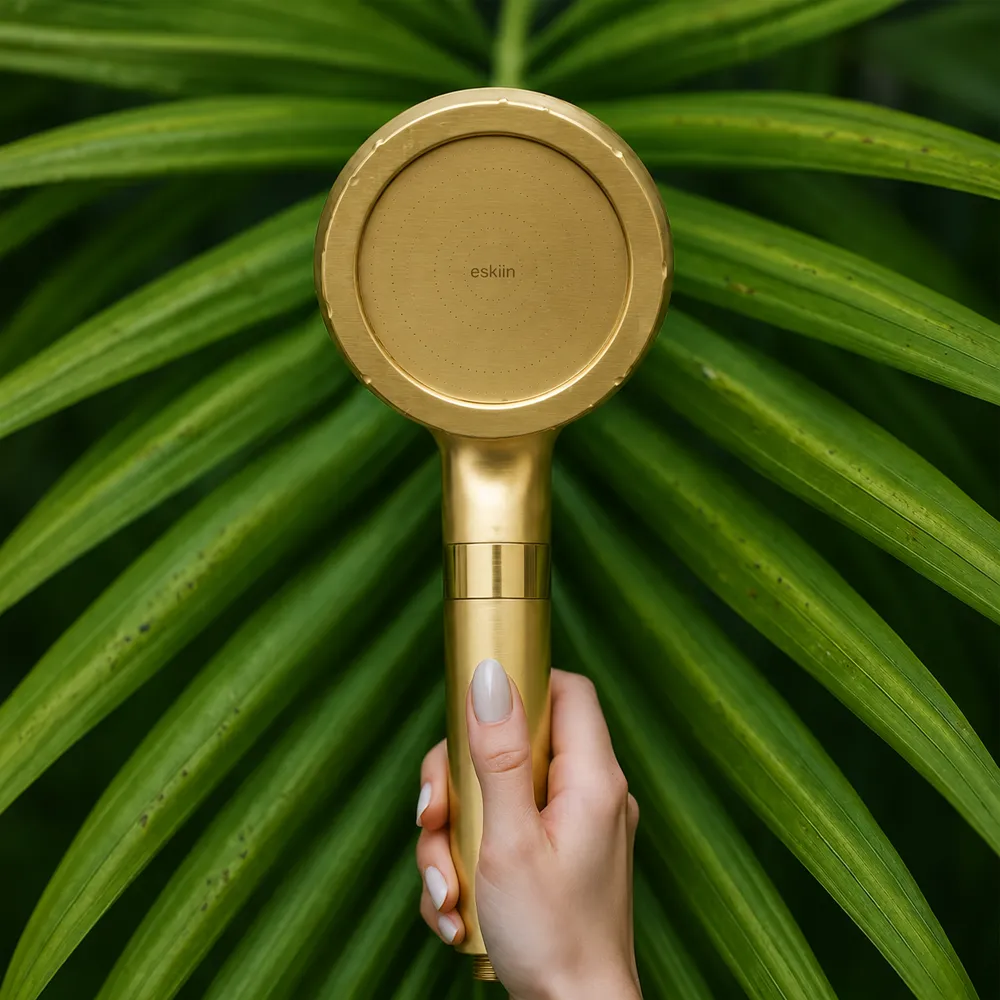 A hand holds a gold-colored shower head in front of a large green palm leaf.