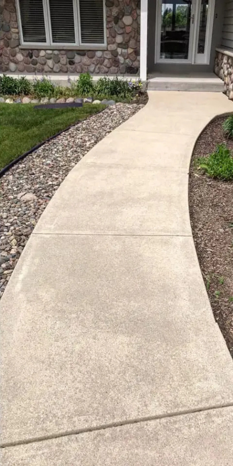 Curved concrete walkway leading to a house entrance with stone facade and garden.