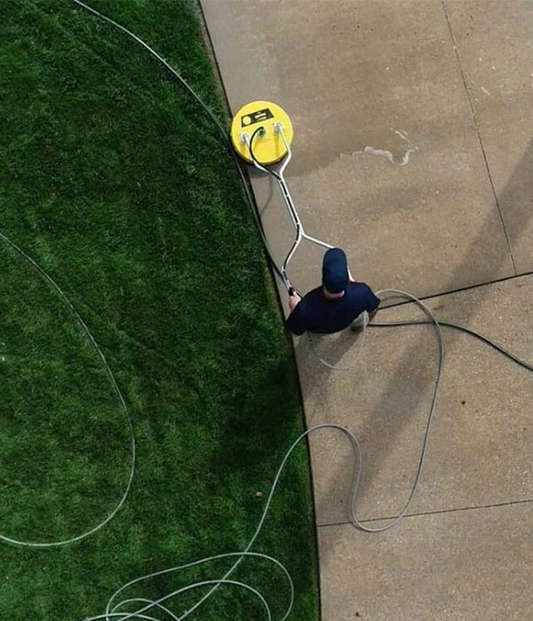 Person using a yellow surface cleaner on a concrete path next to grass.