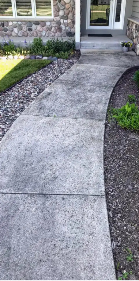 Curved concrete walkway leading to a house entrance with stone facade and plants.