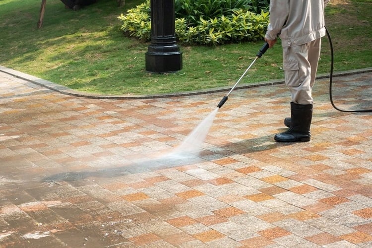 Person pressure washing a tiled outdoor path near grass and plants.