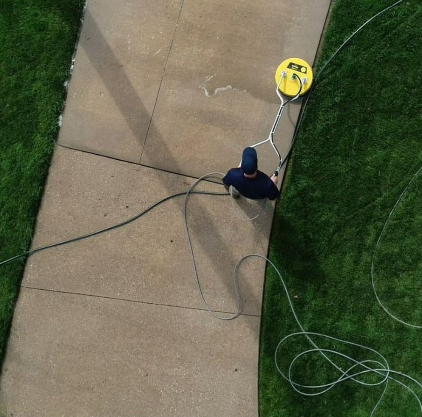Person cleaning a concrete path with a power washer, surrounded by green grass.