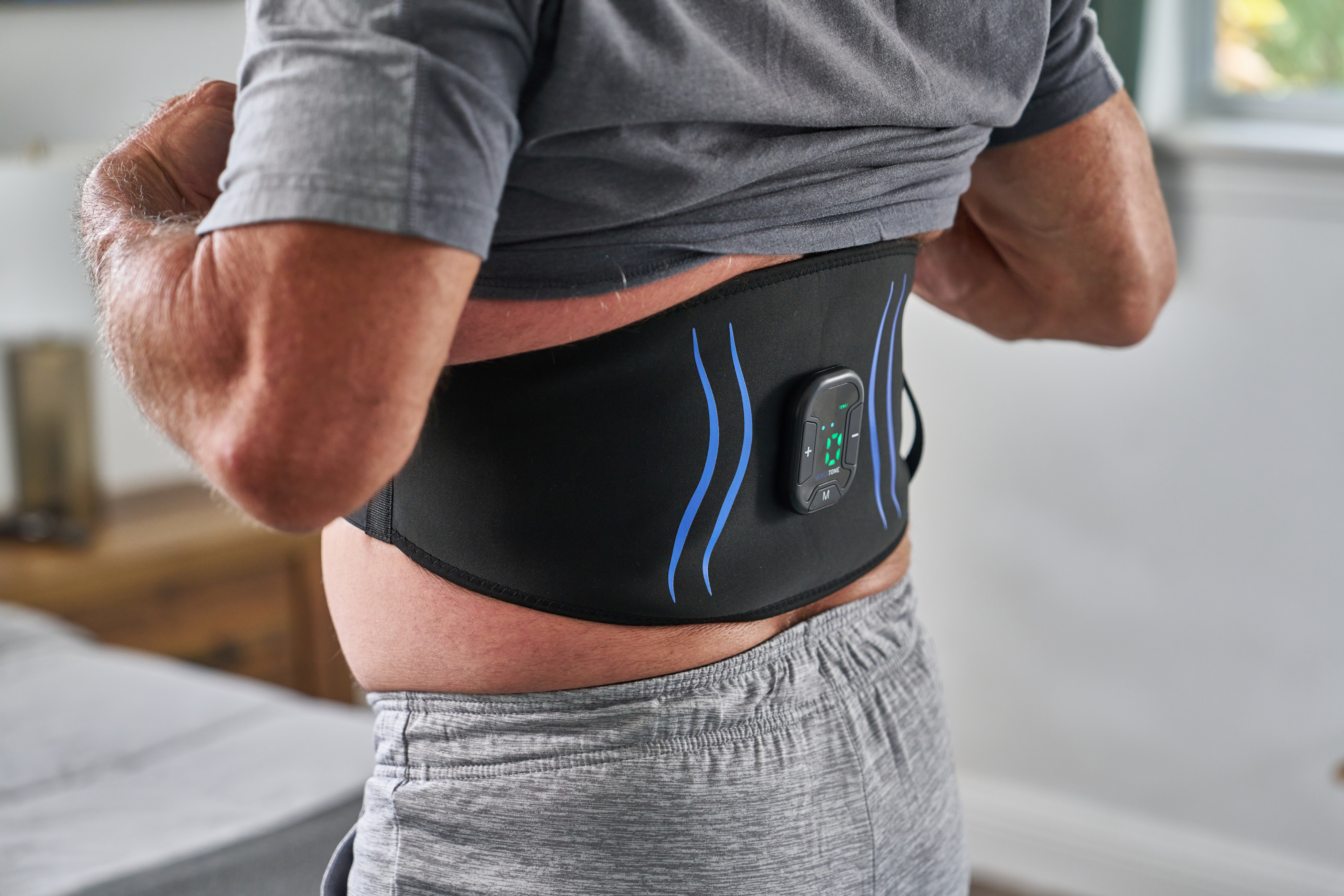 A man from behind lifts his shirt to adjust a black electronic toning belt around his waist.