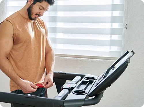 Man adjusting a cord on a treadmill in a sunlit room.