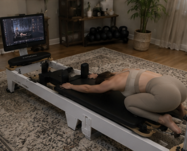 A woman in workout attire stretches on a reformer machine in a home gym setting.