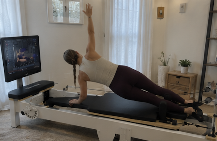 A woman with her back to the camera does a side plank on a Pilates reformer with a screen.