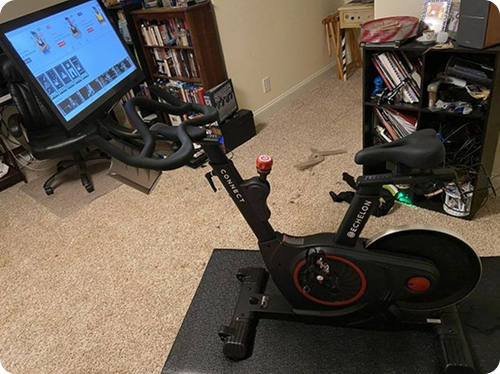 A stationary exercise bike in a room with a screen and shelves in the background.