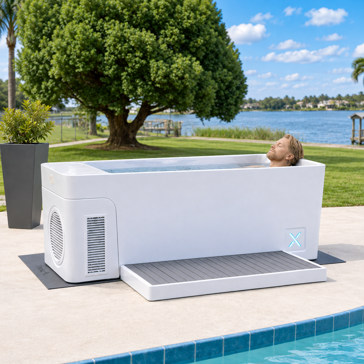 A man with his eyes closed relaxes in a white, modern ice bath tub outdoors by a lake.
