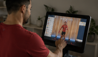 A man in a red shirt touches the screen of an Echelon exercise machine showing a workout class.