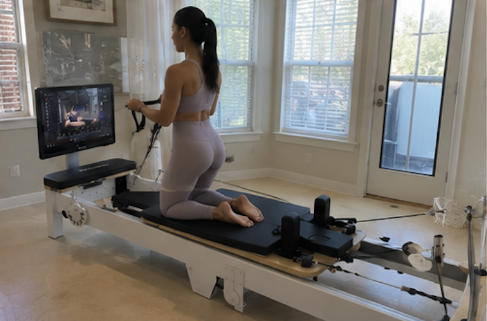 A woman in athletic wear kneels on a Pilates reformer, following a workout on an attached screen.