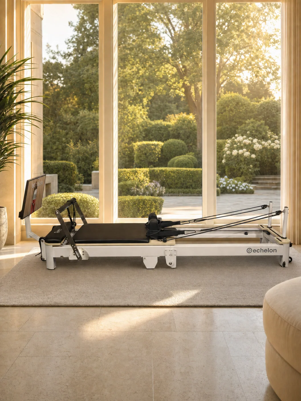 A white Pilates reformer machine in a sunlit room with large windows overlooking a green garden.