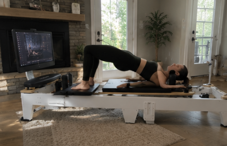 A woman performs a bridge pose on a Pilates reformer machine with a screen in a living room.