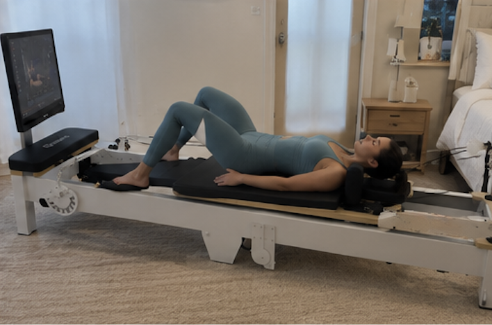 A woman in a blue-green bodysuit lies on a smart pilates reformer machine in a bedroom.