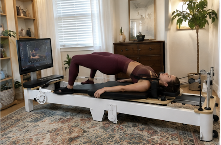 A woman holds a bridge pose on a Pilates reformer with an attached screen in a room.