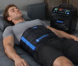 A man lies on his back with a black and blue therapy wrap around his abdomen connected to a machine.