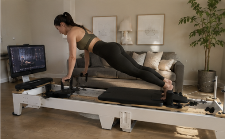 A woman in workout clothes holds a plank position on a Pilates reformer in a living room.