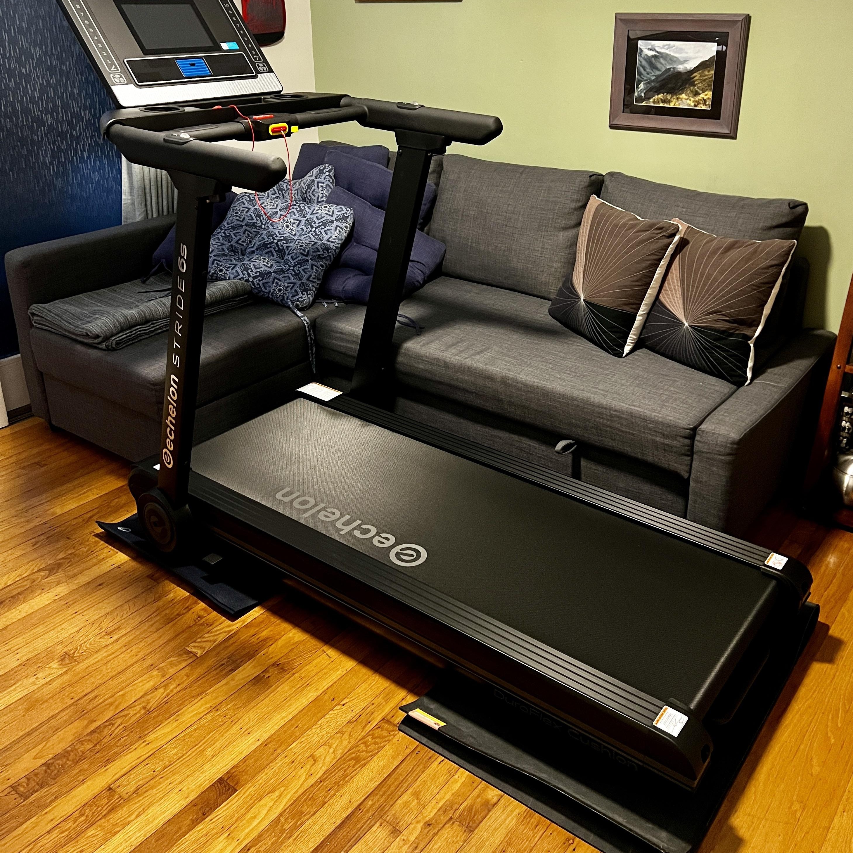 Treadmill in front of a grey couch with cushions in a room with wooden flooring.